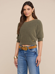 A woman with long brown hair is seen wearing the Guinevere Cropped Pullover in olive green, styled with blue jeans and a tan belt. She stands against a plain light background and completes her look with gold earrings and a gold bracelet.