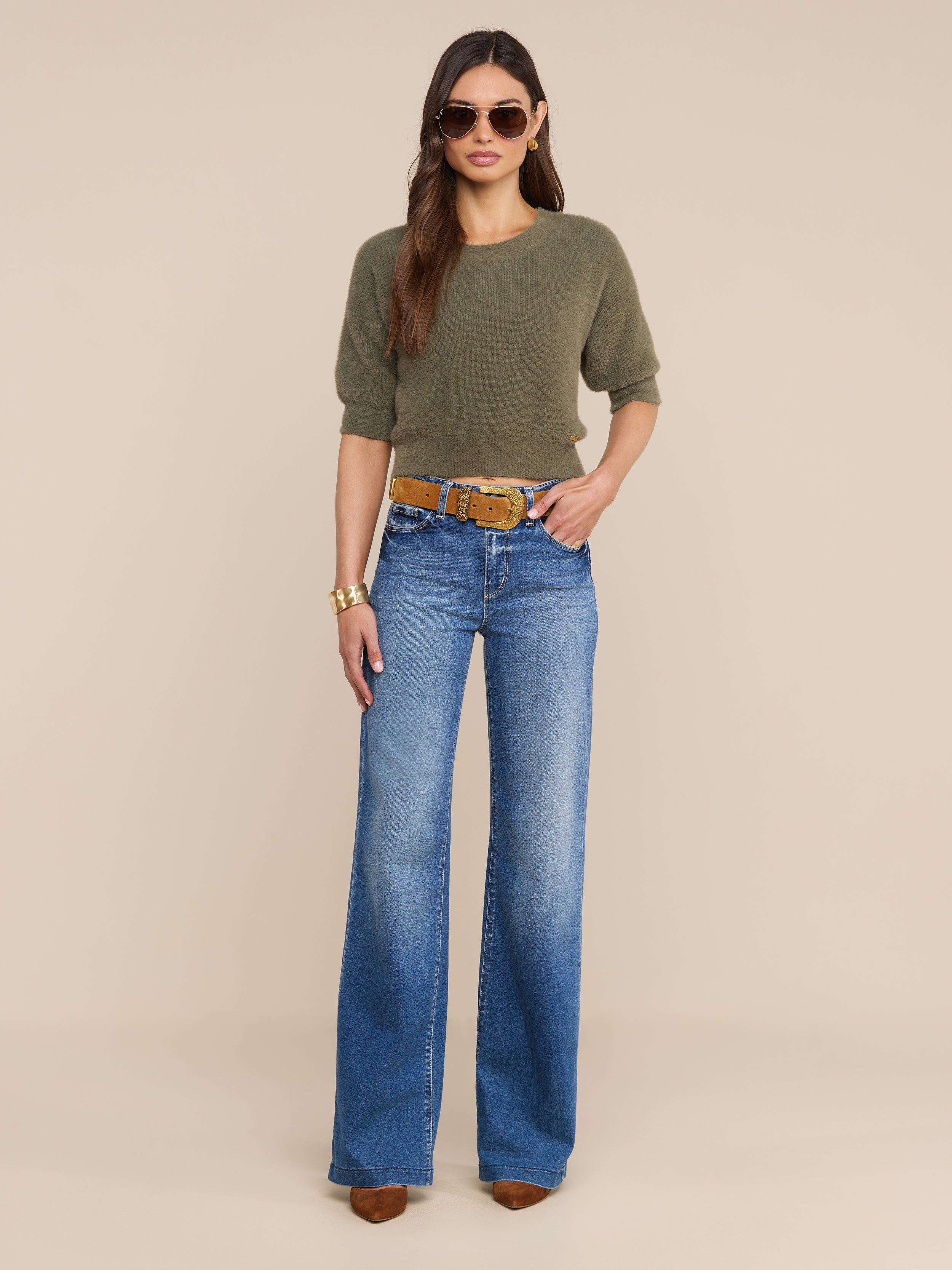 A woman wearing sunglasses, the Guinevere Cropped Pullover in olive green, wide-leg blue jeans with a belt, brown pointed-toe shoes, and a gold bracelet stands against a plain white background.