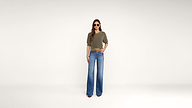 A woman stands against a plain white background, wearing sunglasses, an olive green top, blue wide-leg jeans, brown shoes, and a brown belt. She has long hair and her hands rest at her sides.