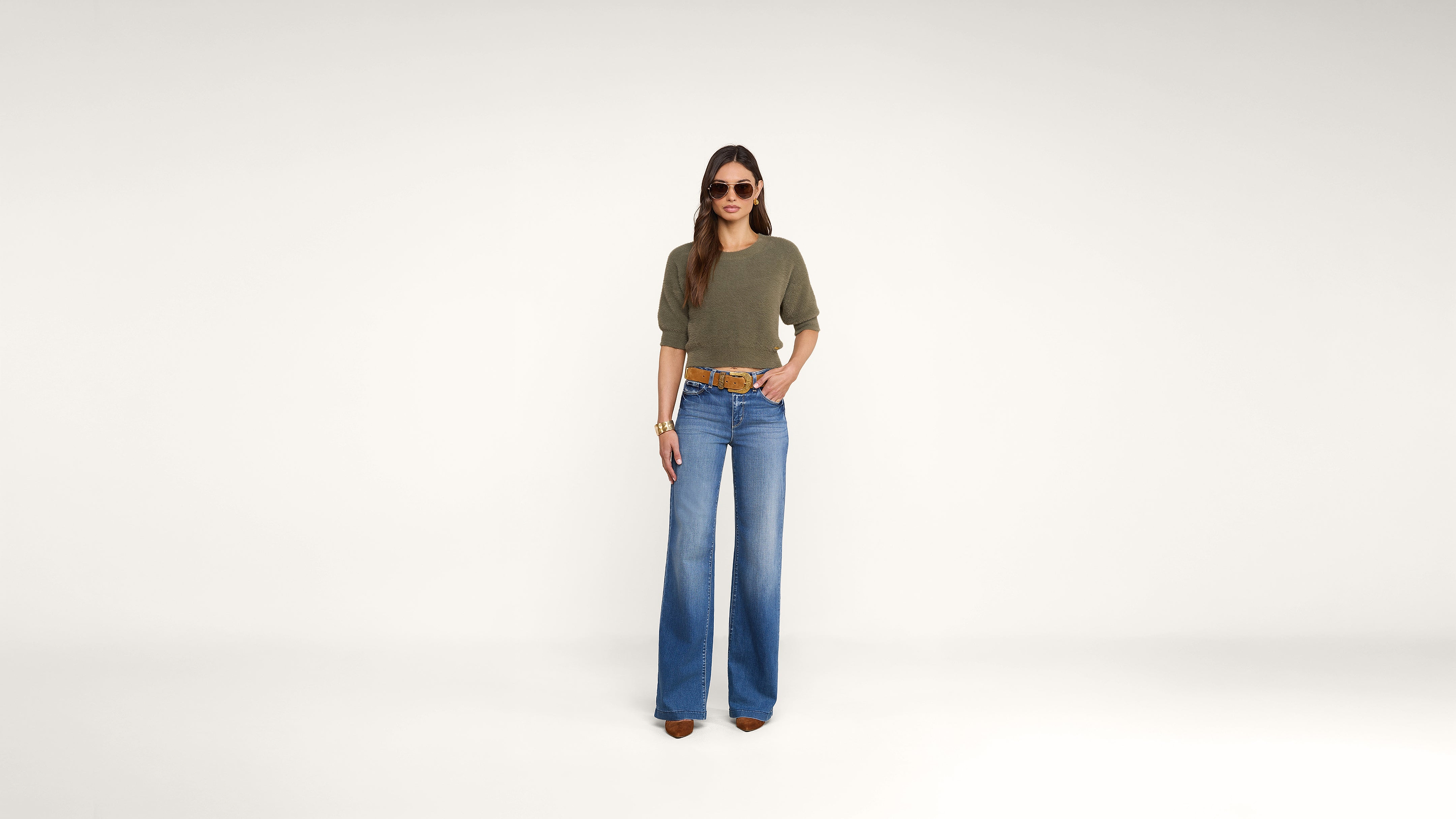 A woman stands against a plain white background, wearing sunglasses, an olive green top, blue wide-leg jeans, brown shoes, and a brown belt. She has long hair and her hands rest at her sides.