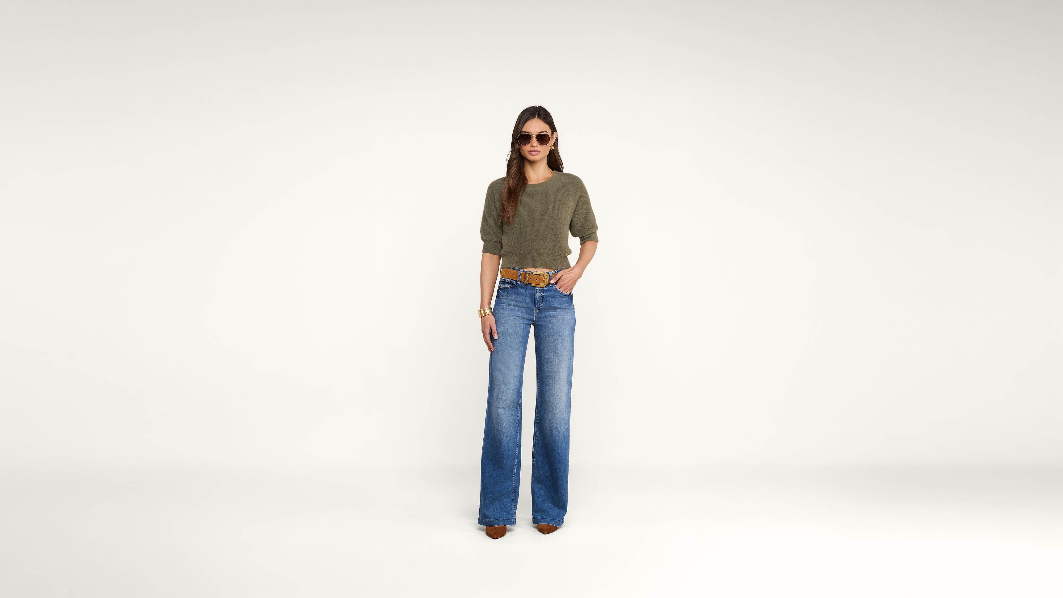 A woman stands against a plain white background, wearing sunglasses, an olive green top, blue wide-leg jeans, brown shoes, and a brown belt. She has long hair and her hands rest at her sides.