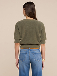 A woman stands facing away, wearing the Guinevere Cropped Pullover in olive green with blue jeans and a tan belt. She has straight brown hair and a watch on her left wrist, set against a plain light background.