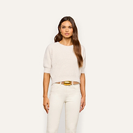 A woman with long brown hair stands facing forward, wearing the Guinevere Cropped Pullover—a white, ultra soft knit crewneck sweater with short sleeves—paired with white pants and a shiny gold belt, against a plain light background.