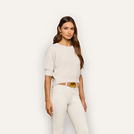 A woman with long brown hair wears the Guinevere Cropped Pullover, an ultra soft knit crewneck sweater, paired with white pants and a tan belt, standing against a plain light background.