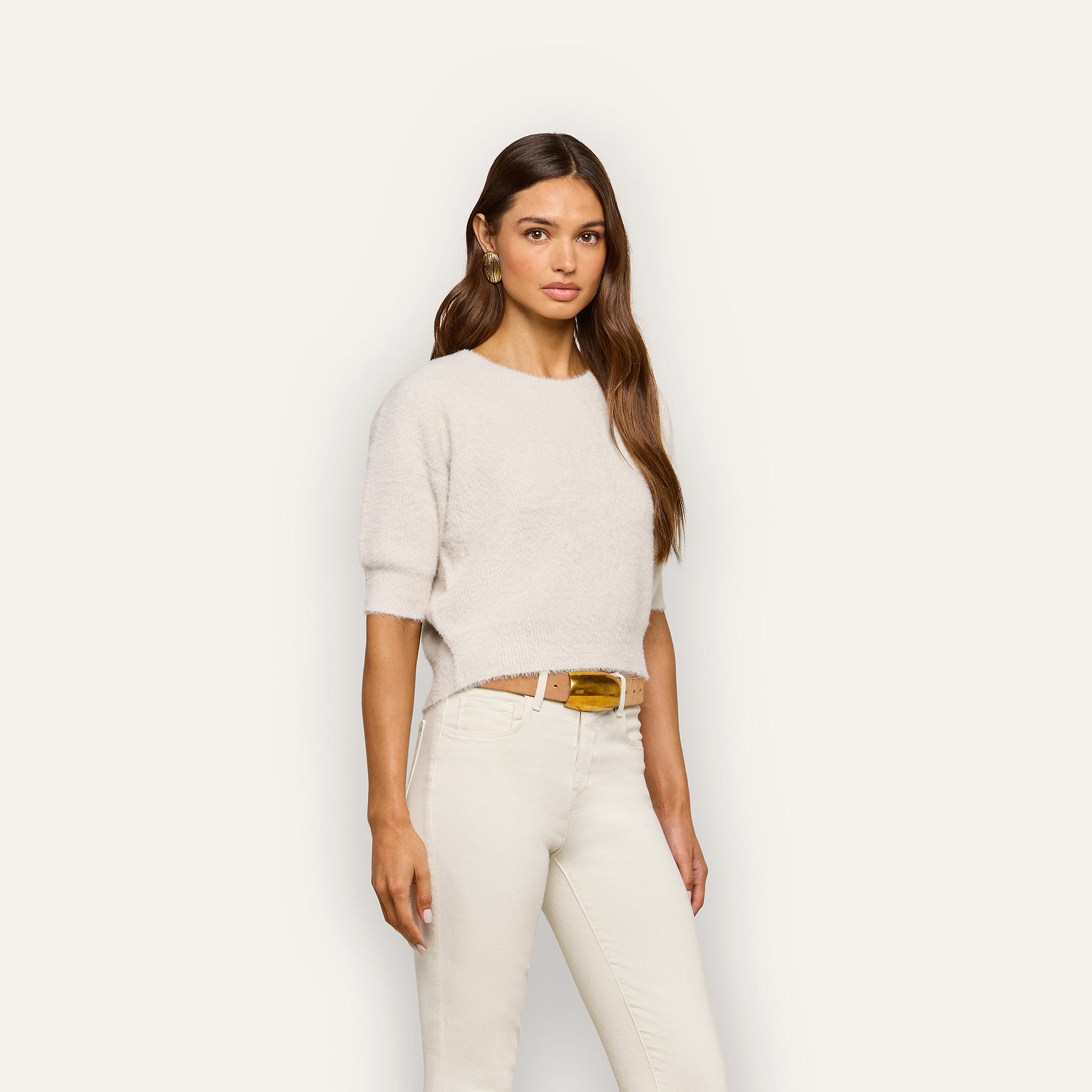 A woman with long brown hair wears the Guinevere Cropped Pullover, an ultra soft knit crewneck sweater, paired with white pants and a tan belt, standing against a plain light background.