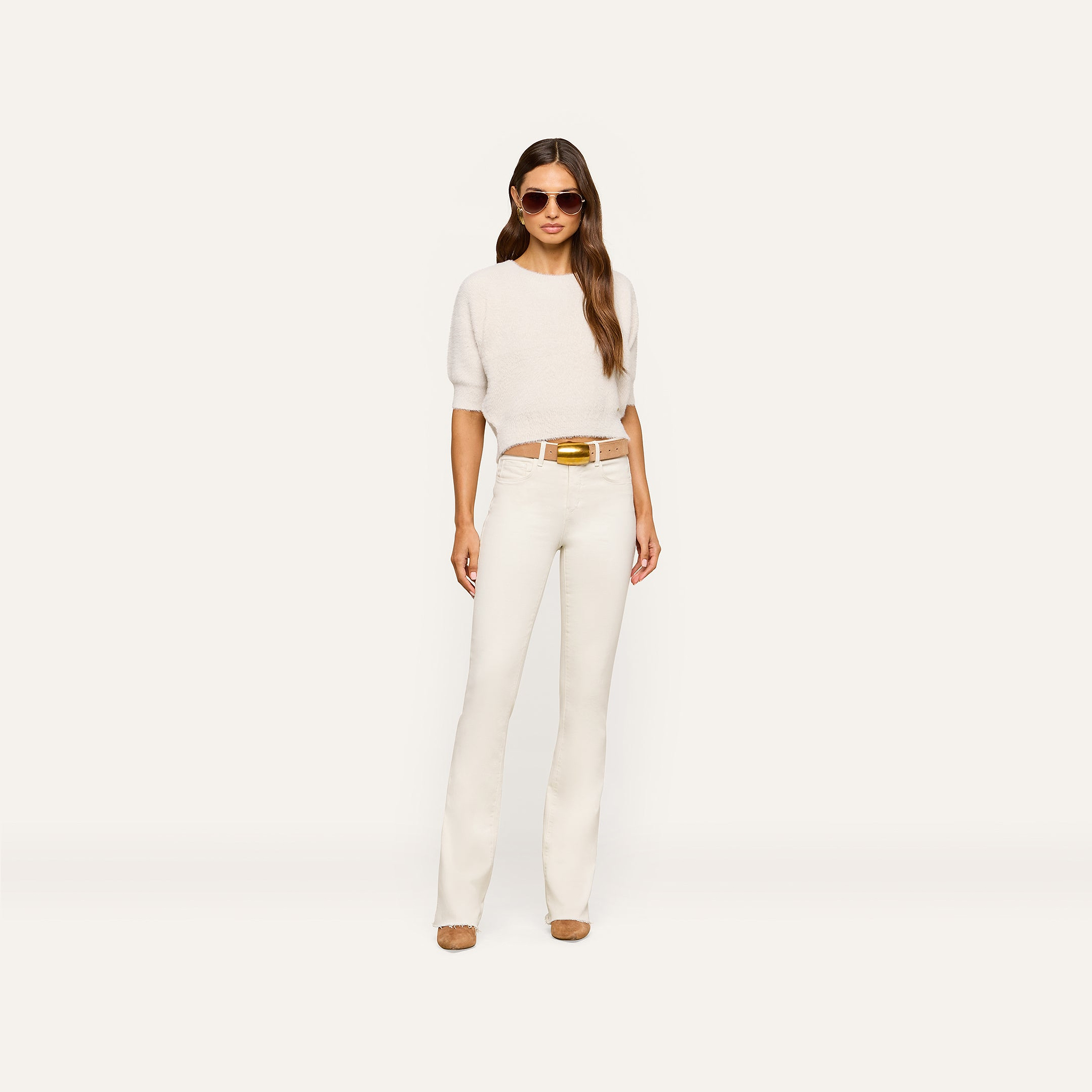 A woman wearing sunglasses, the Guinevere Cropped Pullover—an ultra soft knit crewneck sweater with short sleeves—white pants with a belt, and beige shoes stands against a plain white background.