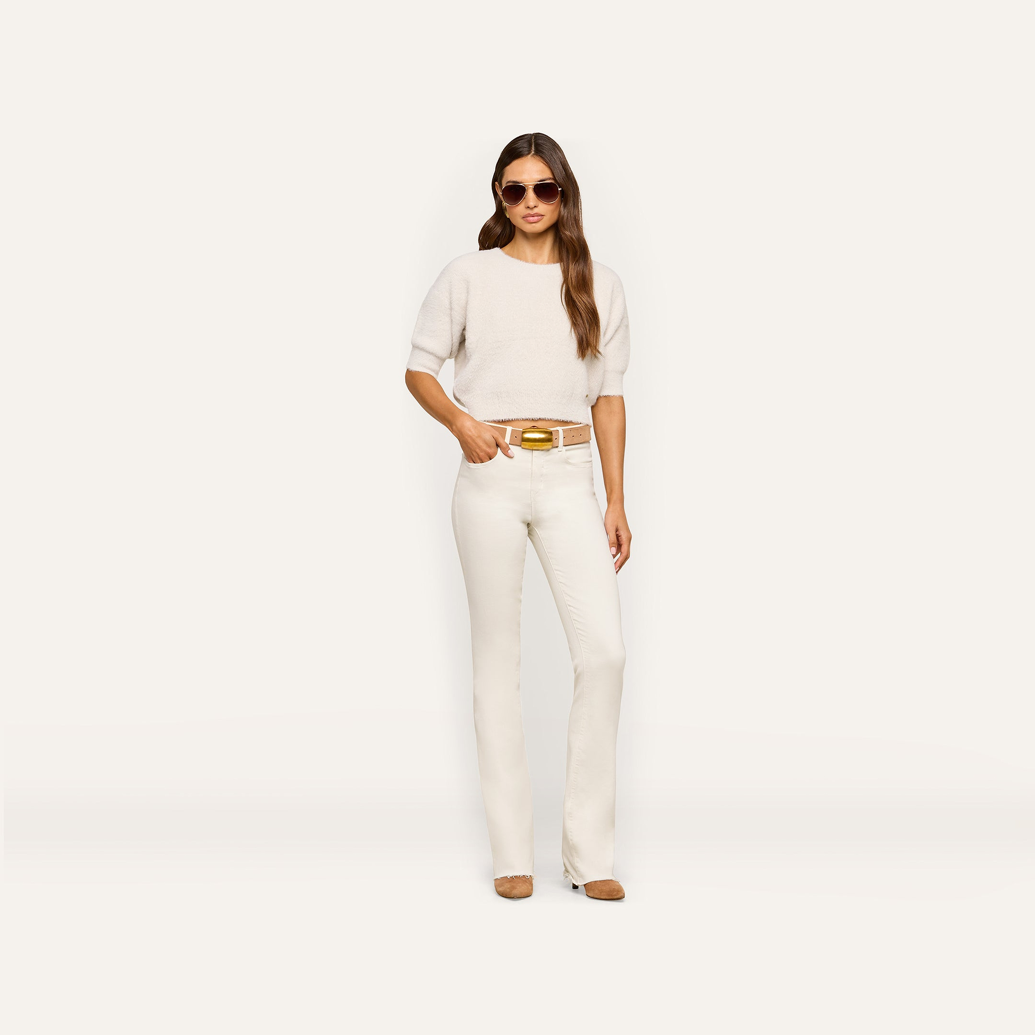 A woman wearing sunglasses, the Guinevere Cropped Pullover in white, white pants, a brown belt, and beige shoes stands against a plain white background with her hand in her pocket.