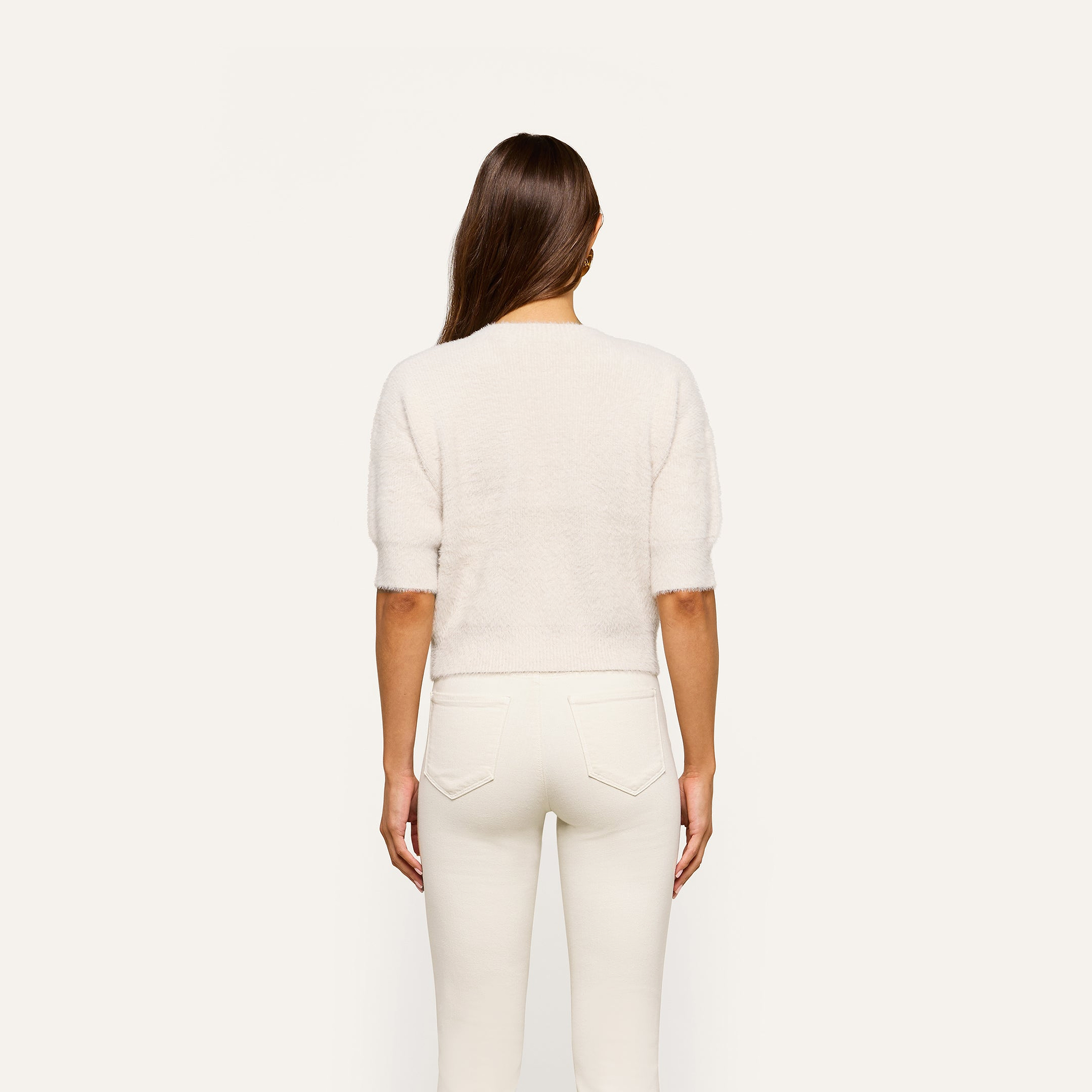 A person with long brown hair stands facing away, wearing the Guinevere Cropped Pullover—an ultra-soft knit crewneck sweater with a cropped silhouette—paired with white jeans against a plain light background.