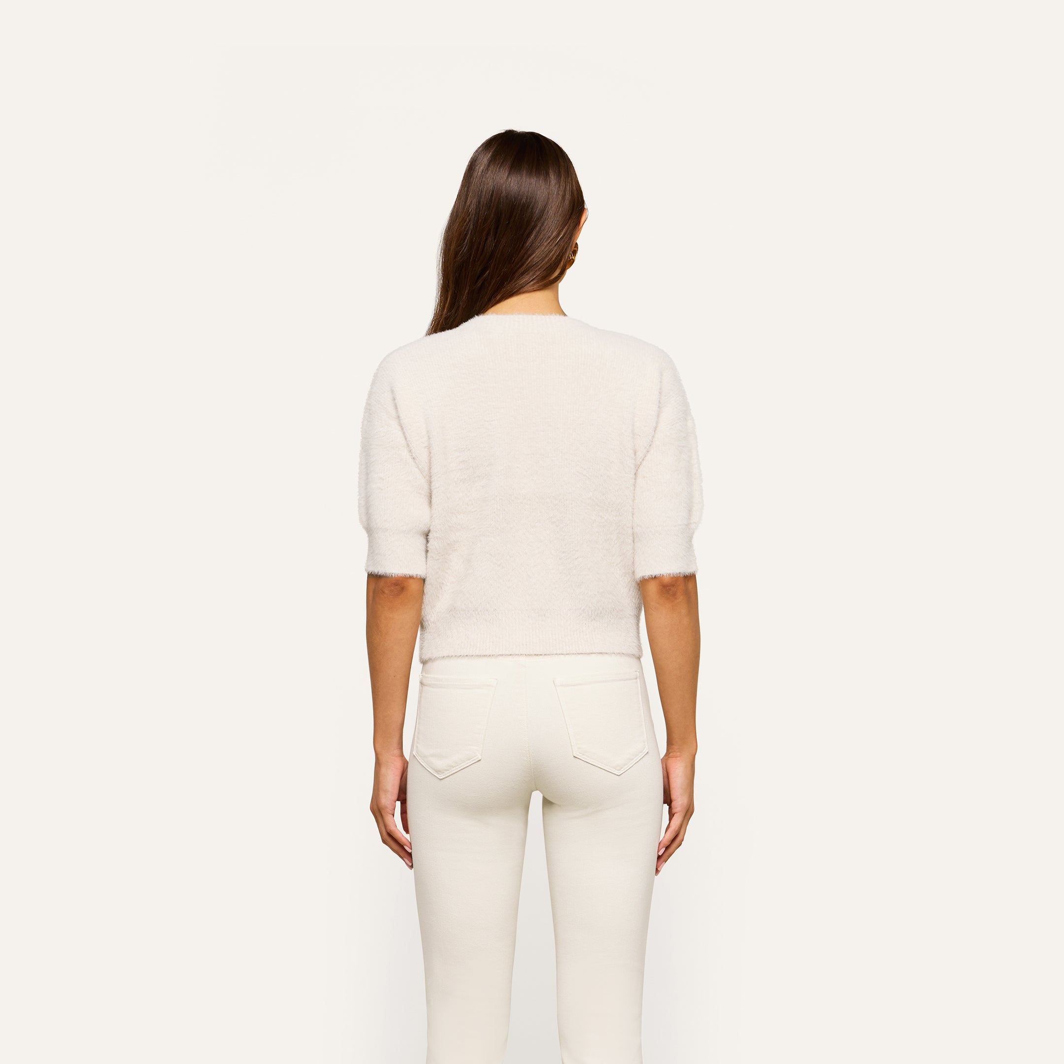 A person with long brown hair stands facing away, wearing the Guinevere Cropped Pullover—an ultra-soft knit crewneck sweater with a cropped silhouette—paired with white jeans against a plain light background.