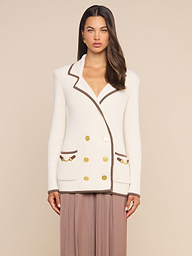 A woman stands against a plain background, wearing the Ranch Knit Blazer—a white double-breasted piece featuring horse bit accents and gold buttons—paired with a light brown skirt.