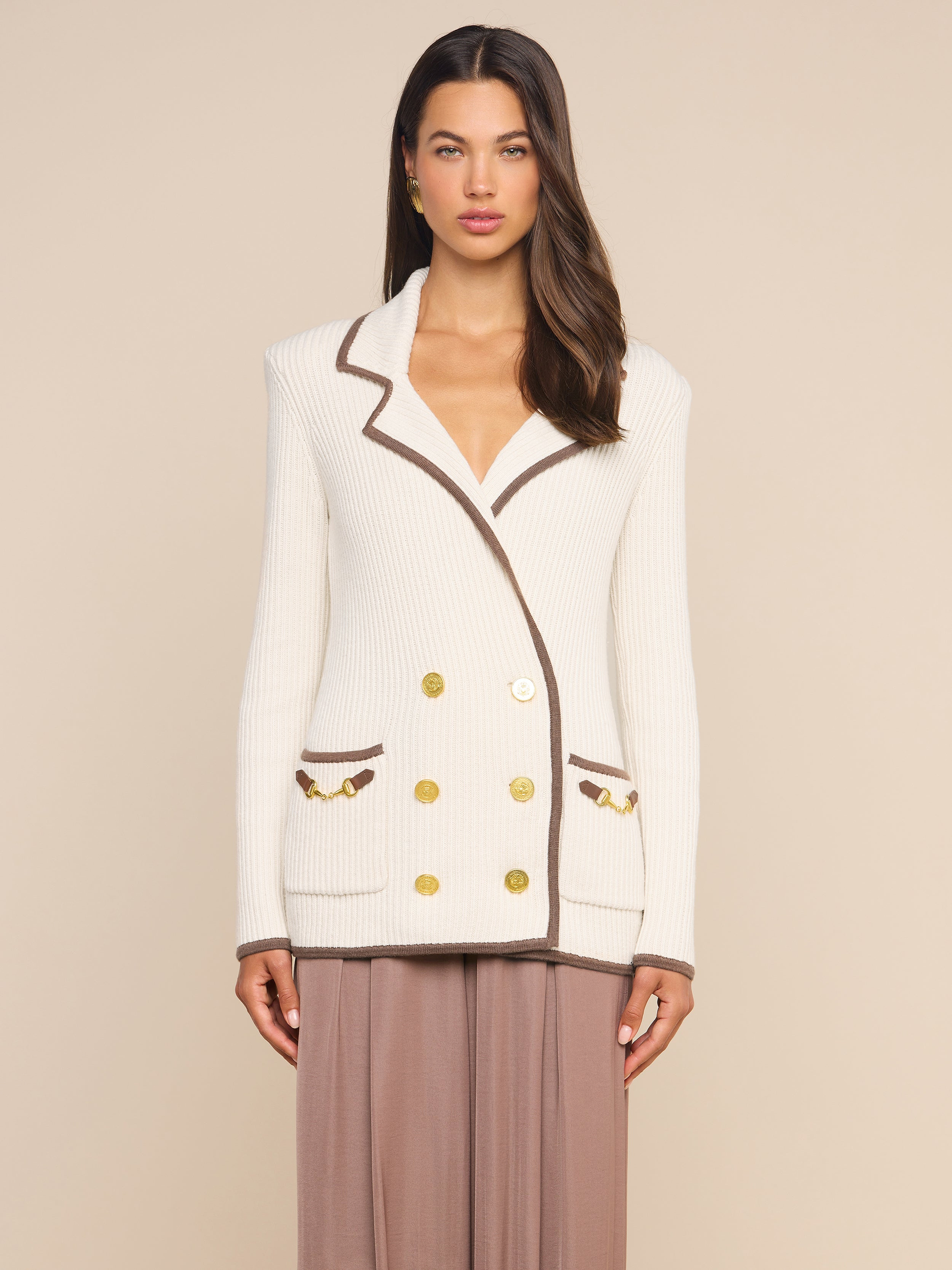 A woman stands against a plain background, wearing the Ranch Knit Blazer—a white double-breasted piece featuring horse bit accents and gold buttons—paired with a light brown skirt.