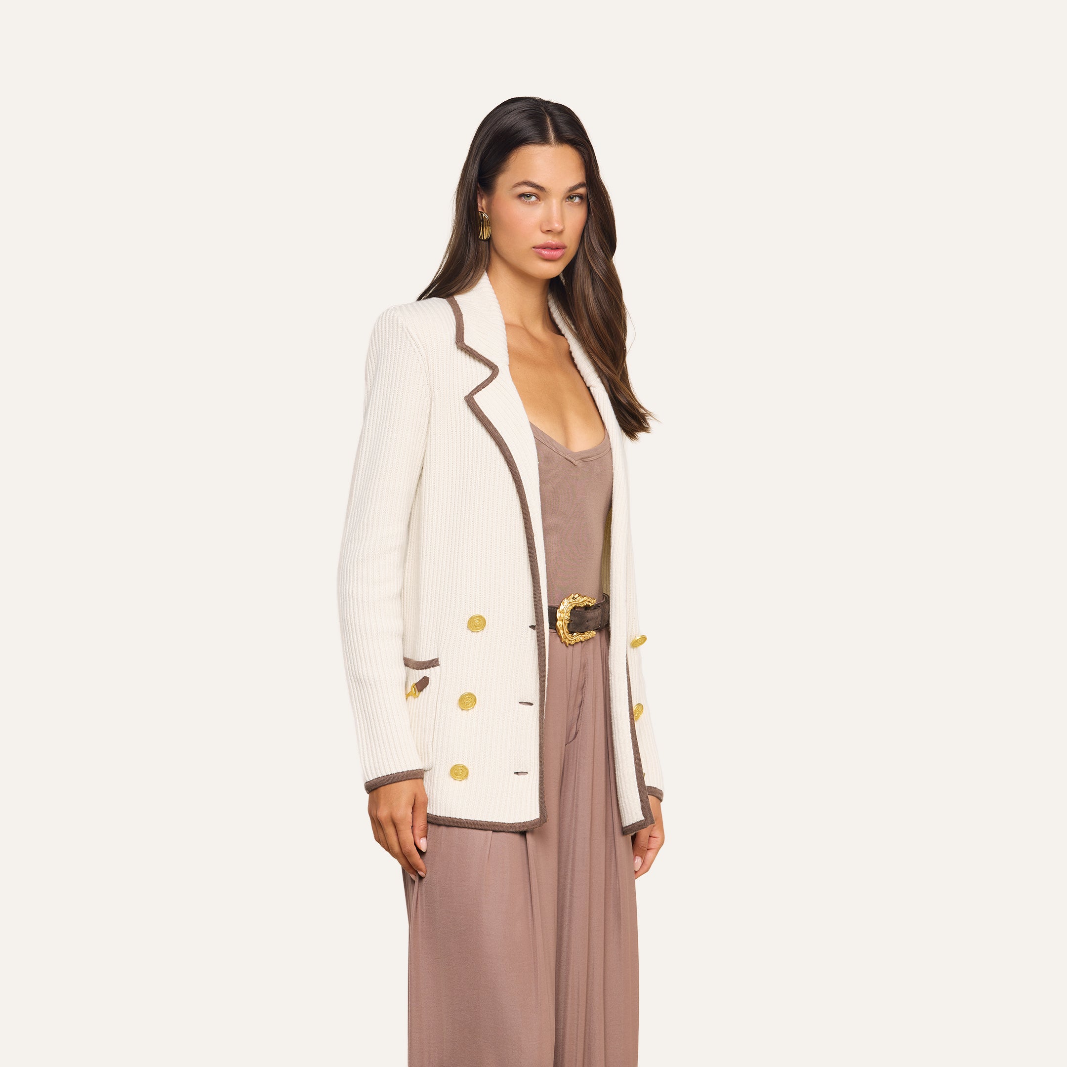 A woman wearing a beige jumpsuit with a wide belt and the Ranch Knit Blazer—a double-breasted style featuring brown trim and gold buttons—stands against a plain, light background.