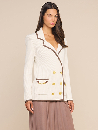 A woman wearing the Ranch Knit Blazer, a white double-breasted piece with gold buttons and brown trim, pairs it with a flowing mauve ribbed knit skirt while standing against a neutral background.