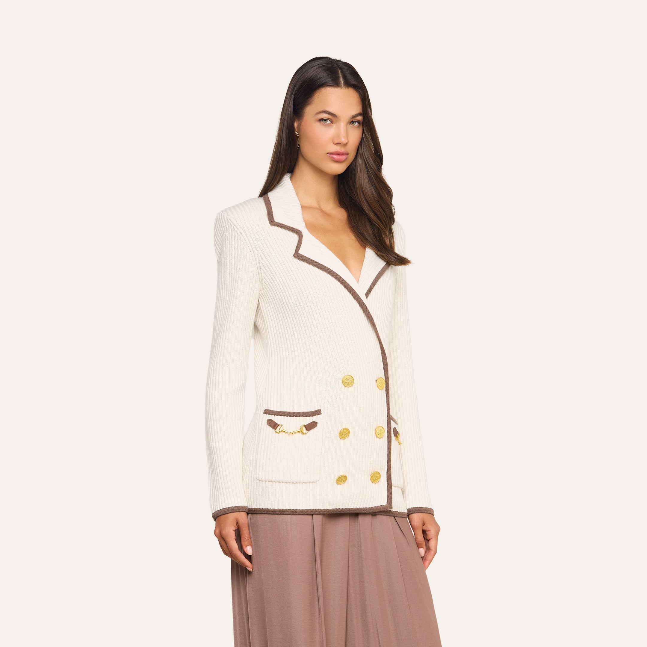 Dressed in the Ranch Knit Blazer—a cream double-breasted piece featuring brown trim, gold buttons, and subtle horse bit accents—a woman pairs it with a flowy taupe skirt while standing against a plain light background.