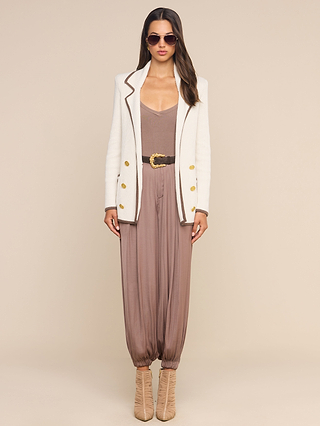 Wearing the Ranch Knit Blazer, a woman in sunglasses stands against a neutral background. The crisp white, double-breasted blazer with gold buttons is styled over a taupe jumpsuit, belted at the waist and paired with tan heeled boots.