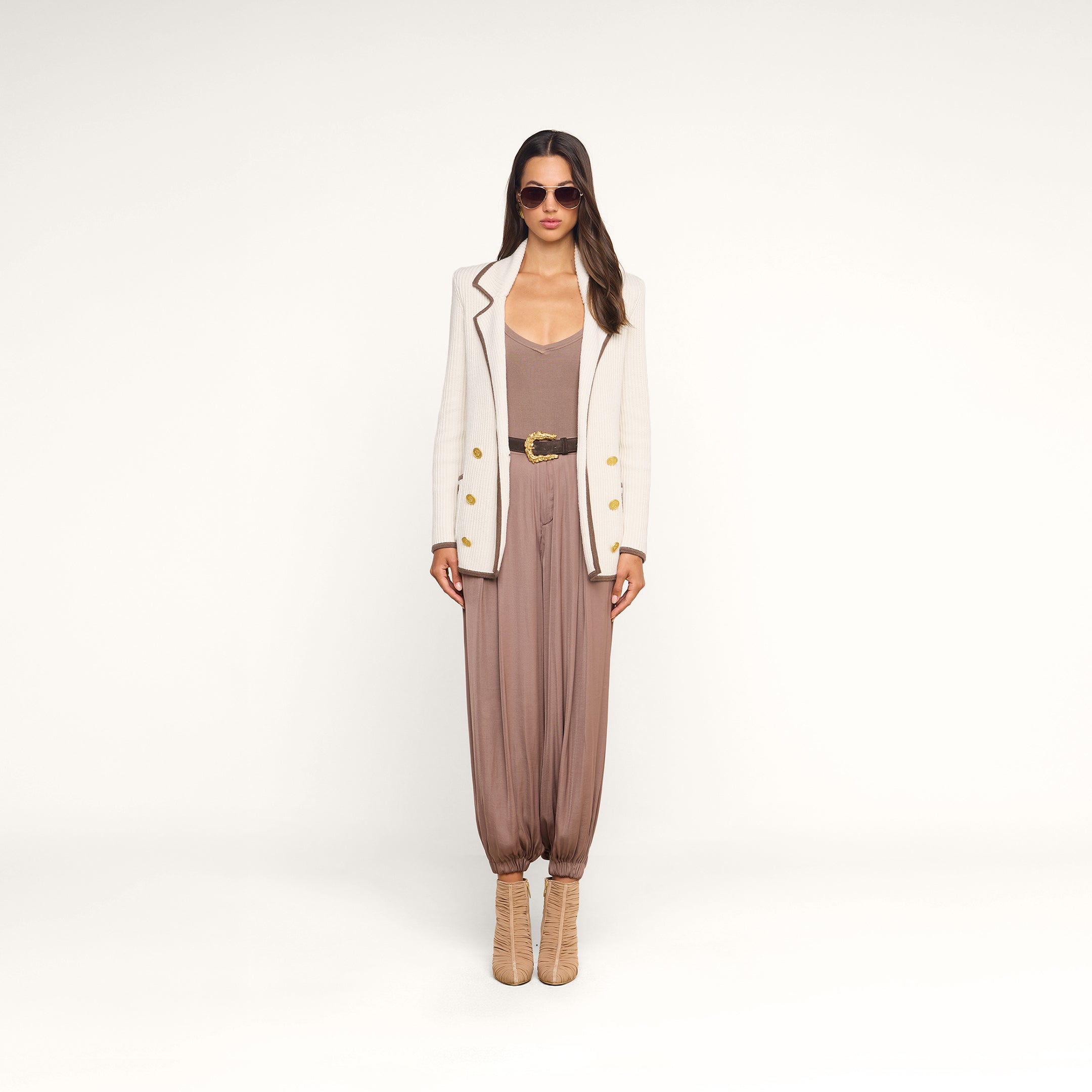 A woman stands against a plain white background wearing sunglasses, tan heeled boots, a taupe jumpsuit with a wide belt, and the Ranch Knit Blazer—a double-breasted white blazer with gold buttons.