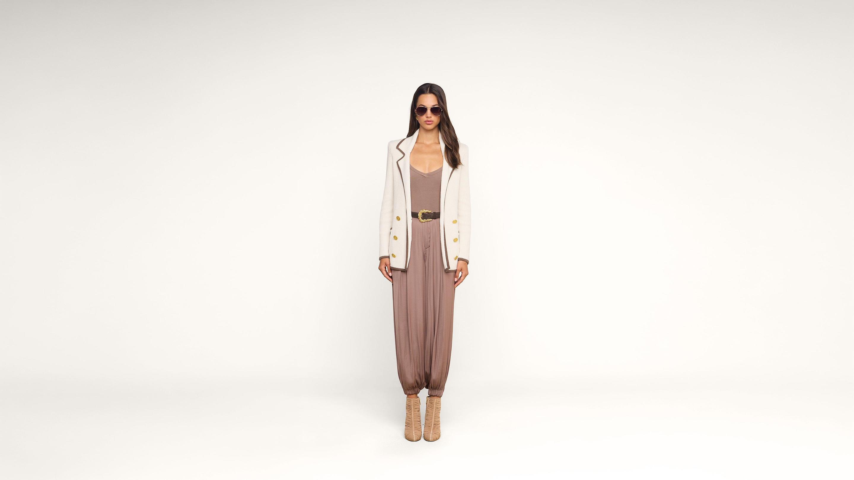 A woman stands against a white background wearing sunglasses, a white blazer, a brown jumpsuit, a wide belt, and tan ankle boots. She faces forward with her arms relaxed at her sides.