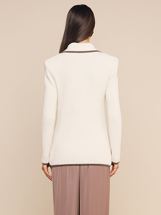 A woman stands with her back to the camera, wearing the Ranch Knit Blazer—a white ribbed knit long-sleeve sweater with brown trim—paired with loose taupe pants, set against a plain beige background.