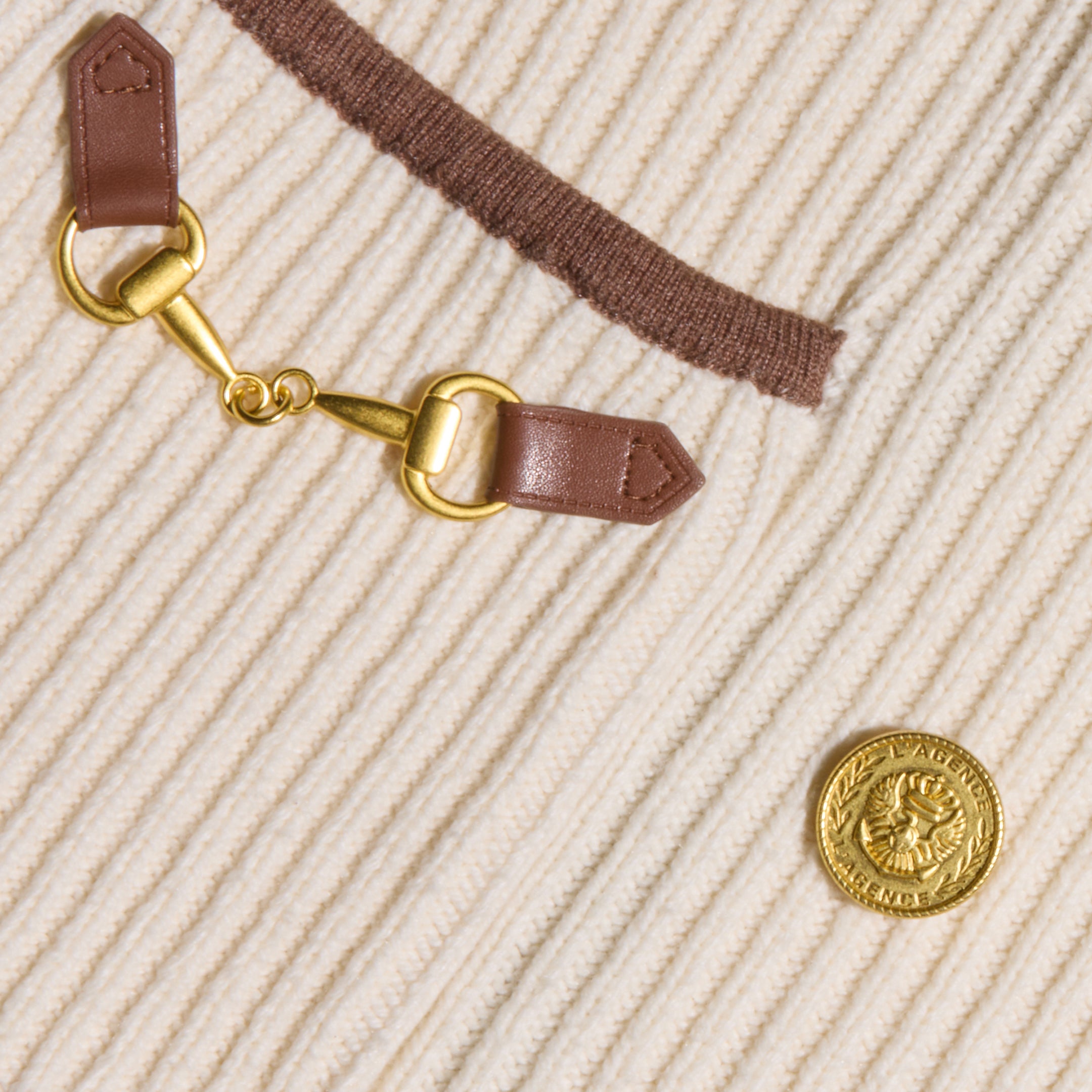 Close-up of the Ranch Knit Blazer, featuring cream-colored ribbed knit fabric with brown trim, a gold emblem button, and a gold clasp connecting two brown leather straps.