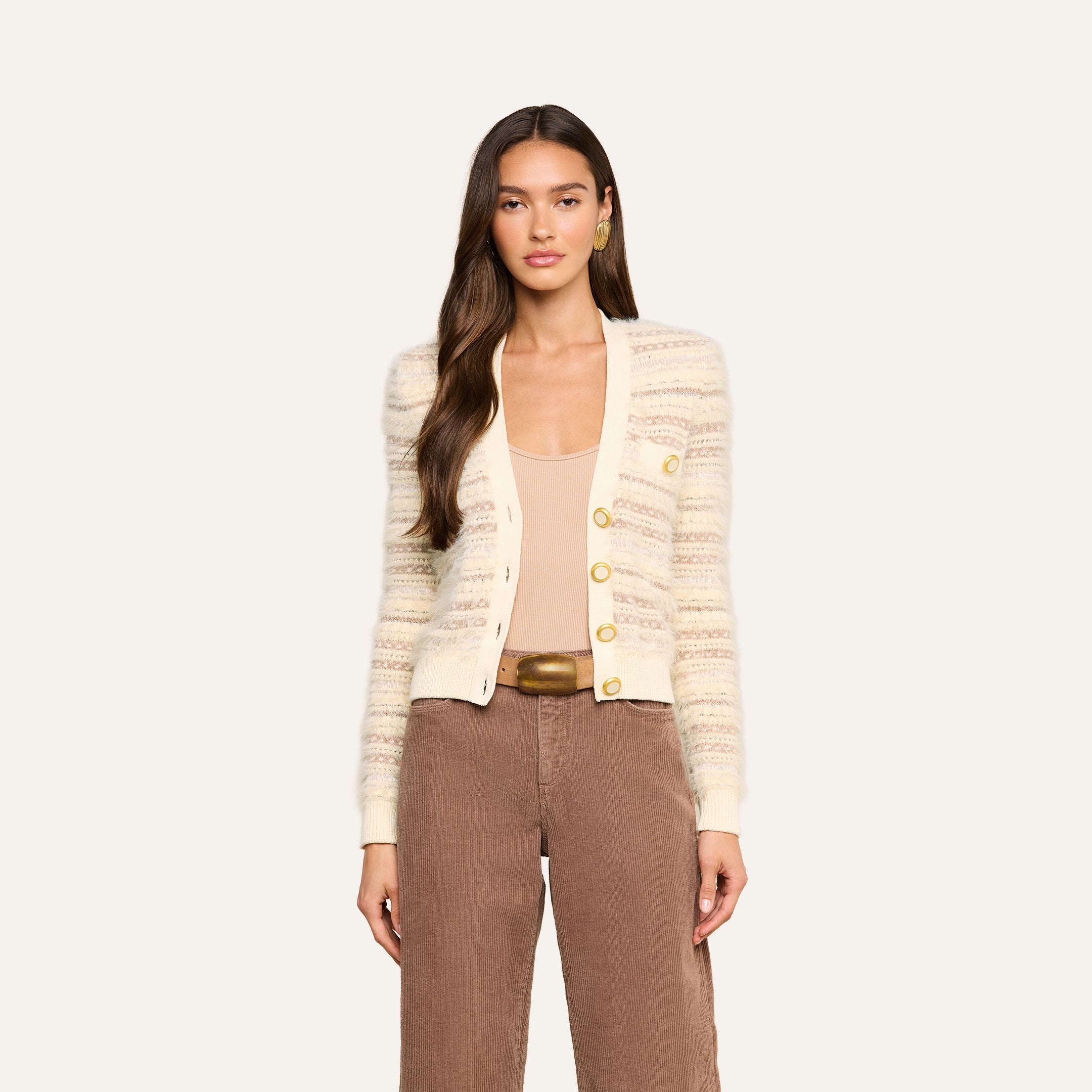 Woman with long brown hair wearing the Ariadne Cardigan, a cream striped piece crafted from cotton-blend yarns with gold buttons, styled with a beige top, brown high-waisted pants, and gold hoop earrings, standing against a plain light background.