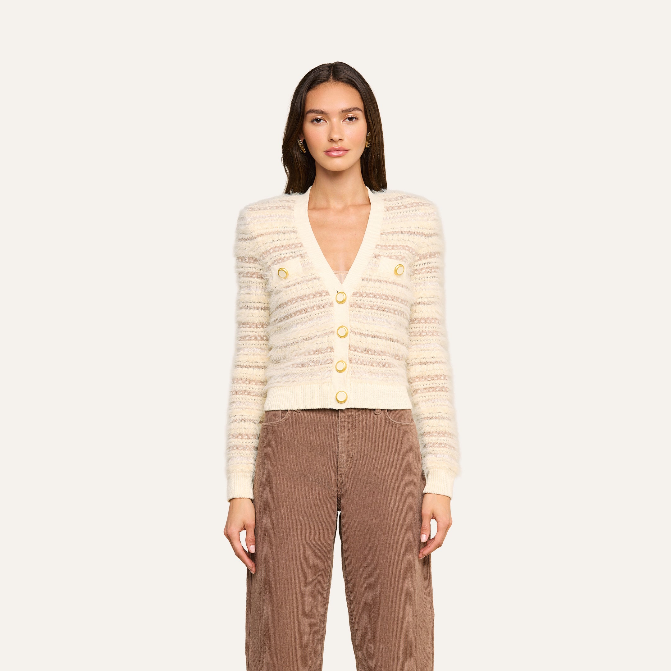 A woman stands against a plain light background, facing forward with a neutral expression, wearing the Ariadne Cardigan—a cream and beige striped piece crafted from cotton-blend mixed stitches with gold buttons—and brown pants.