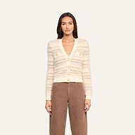 A woman stands against a plain light background, facing forward with a neutral expression, wearing the Ariadne Cardigan—a cream and beige striped piece crafted from cotton-blend mixed stitches with gold buttons—and brown pants.
