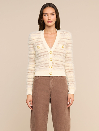 A woman stands against a neutral background wearing the Ariadne Cardigan, a cream and beige striped fuzzy cardigan crafted from cotton-blend mixed stitches with gold buttons, paired with brown corduroy pants.
