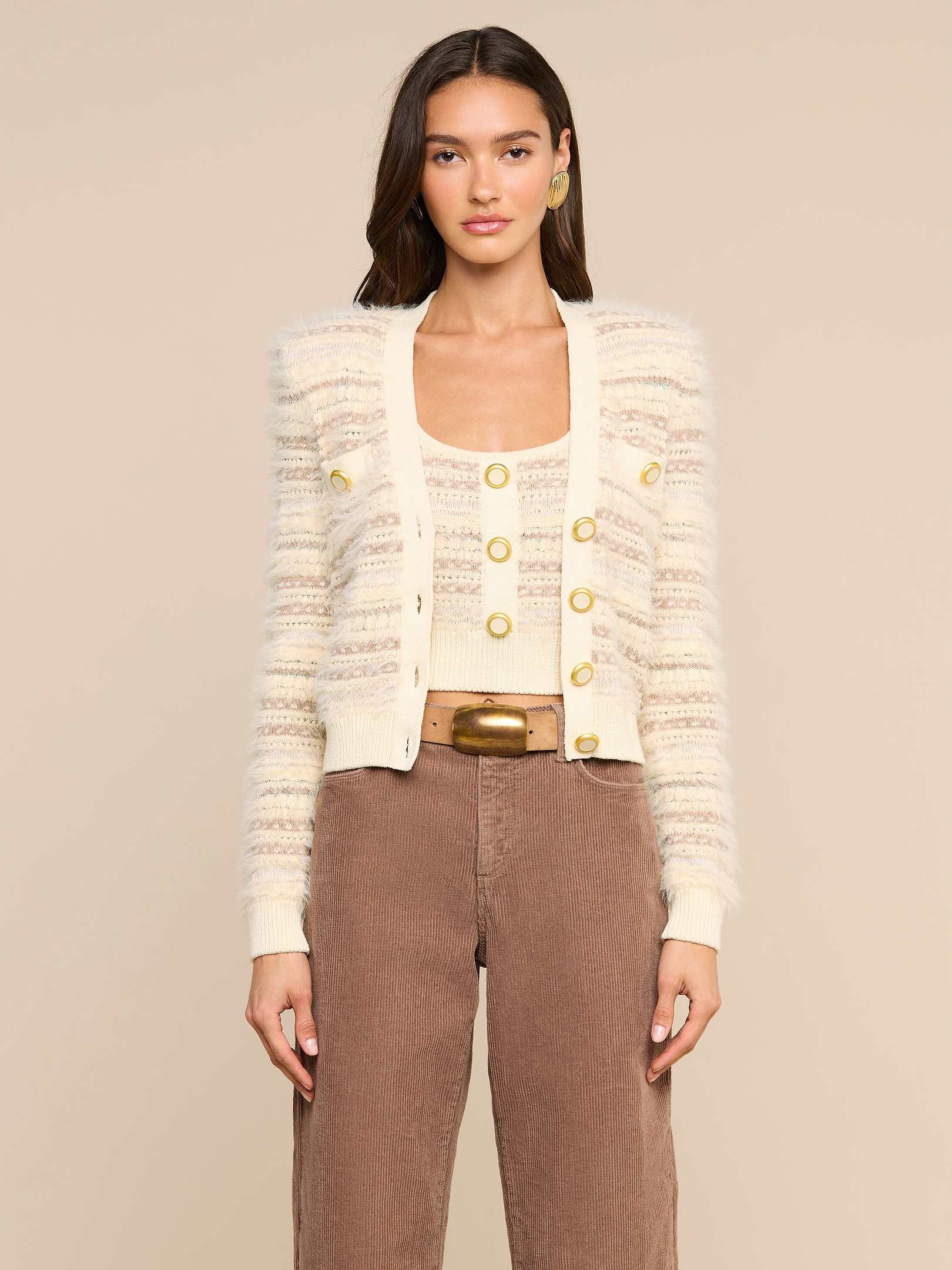 Wearing the Ariadne Cardigan—a cream and beige striped piece crafted from a cotton-blend with mixed stitches and gold buttons—layered over a matching top, brown high-waisted pants, and accessorized with gold hoop earrings, a woman stands against a plain light background.