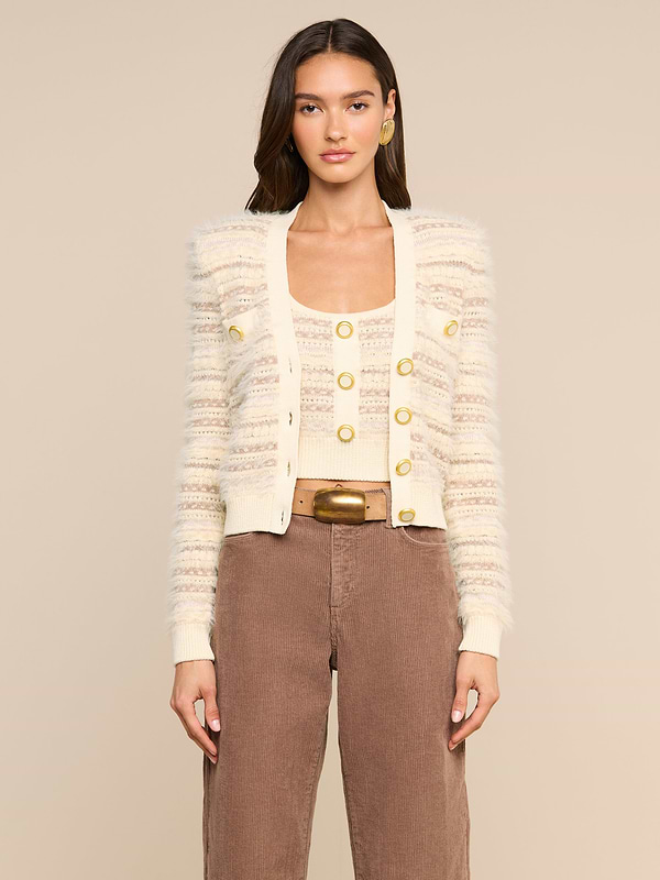 Wearing the Ariadne Cardigan—a cream and beige striped piece crafted from a cotton-blend with mixed stitches and gold buttons—layered over a matching top, brown high-waisted pants, and accessorized with gold hoop earrings, a woman stands against a plain light background.