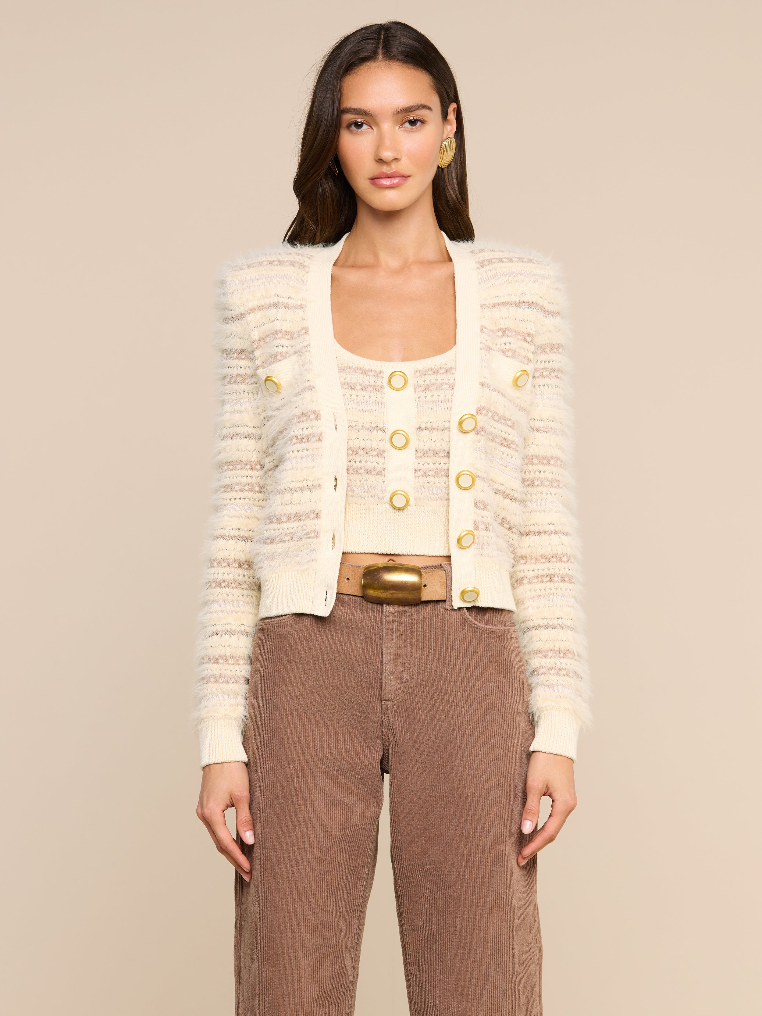 Wearing the Ariadne Cardigan—a cream and beige striped piece crafted from a cotton-blend with mixed stitches and gold buttons—layered over a matching top, brown high-waisted pants, and accessorized with gold hoop earrings, a woman stands against a plain light background.