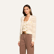Wearing the Ariadne Cardigan, a cream and beige striped design crafted from cotton-blend mixed stitches, paired with a matching top and brown pants, a woman poses against a plain light background.