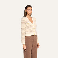 A woman wearing the Ariadne Cardigan—crafted from a cream and beige cotton-blend with gold buttons—paired with brown corduroy pants stands against a plain light background.