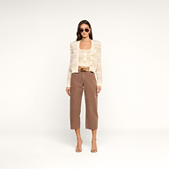 A woman with straight hair and a neutral expression stands against a plain white background wearing sunglasses, the Ariadne Cardigan—a cream striped cardigan crafted from cotton-blend eyelash yarns—over a matching top, brown cropped pants, and open-toe heels.