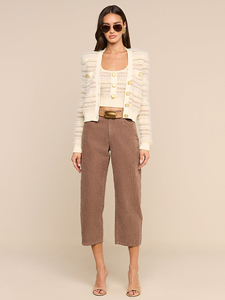 A woman wearing sunglasses and the Ariadne Cardigan—a cream and gold textured piece crafted from mixed stitches and cotton-blend eyelash yarns—paired with a matching top, brown cropped pants, a brown belt, and beige heeled sandals, stands against a plain beige background.