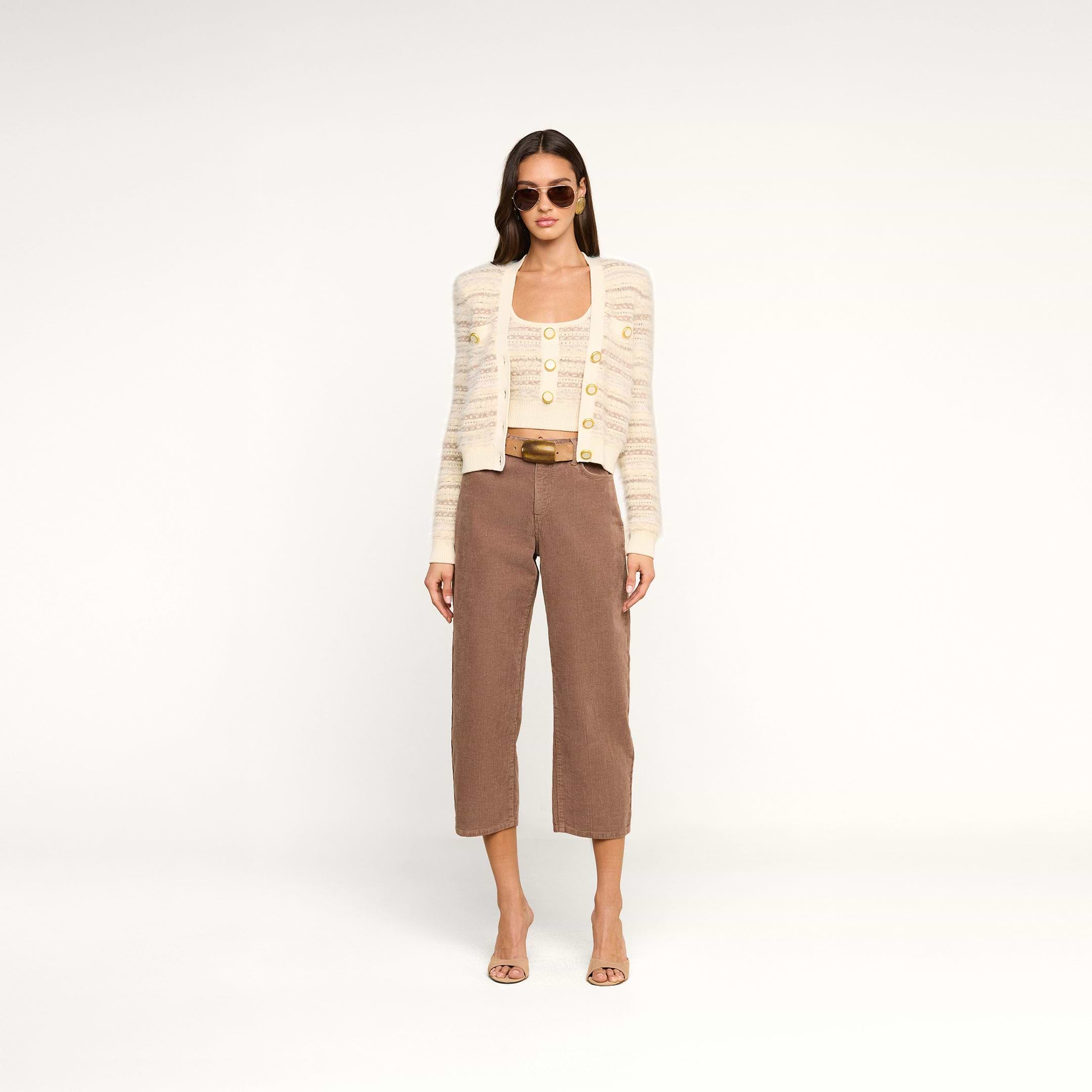 A woman with straight hair and a neutral expression stands against a plain white background wearing sunglasses, the Ariadne Cardigan—a cream striped cardigan crafted from cotton-blend eyelash yarns—over a matching top, brown cropped pants, and open-toe heels.