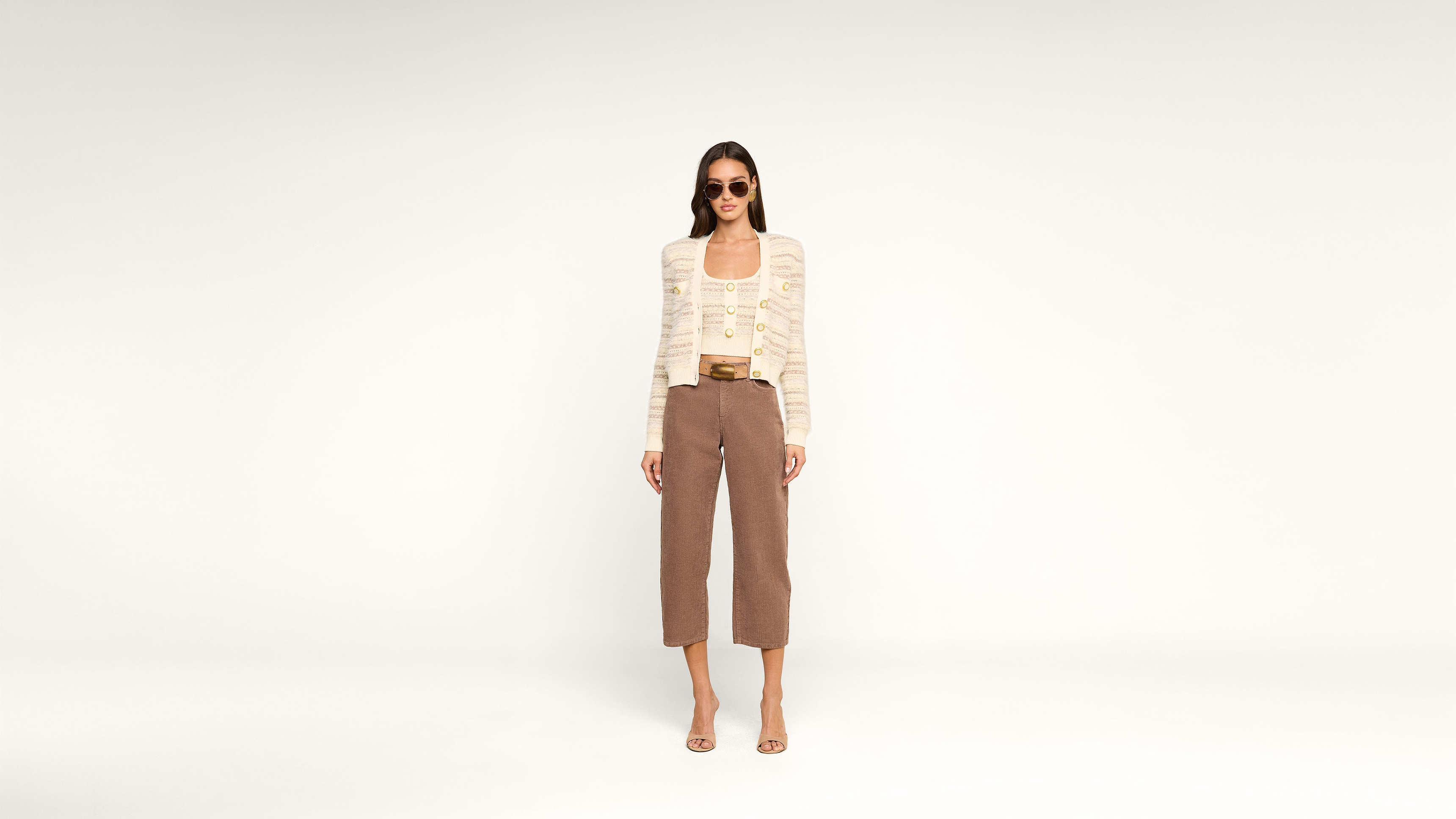 A woman wearing sunglasses, a light knit cardigan, a beige top, cropped brown pants, and heeled sandals stands facing forward in a spacious white studio.