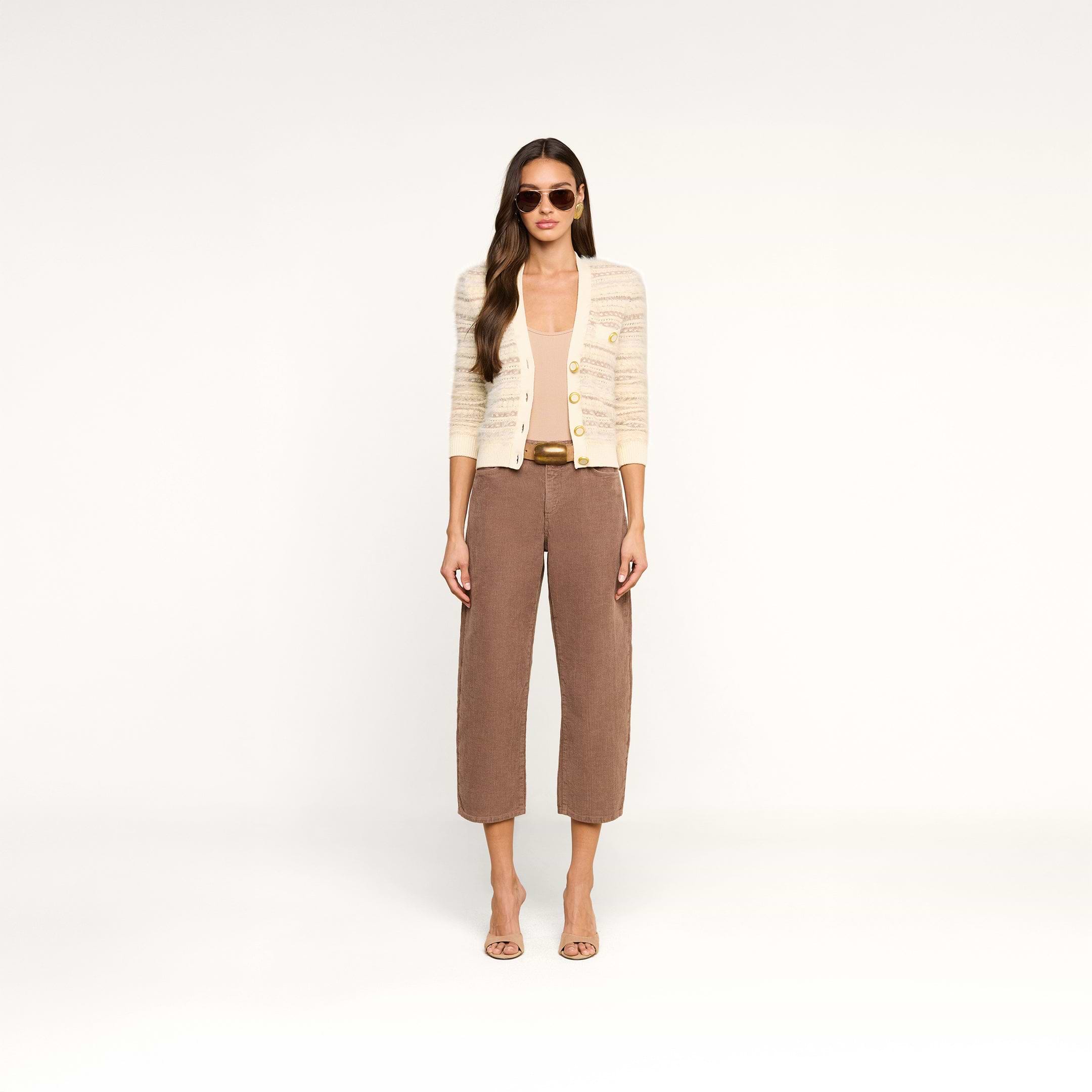 Wearing the Ariadne Cardigan, a woman with long wavy hair stands against a white background. She wears sunglasses, a white top, brown cropped pants with a belt, and open-toe heels, showcasing a neutral expression.