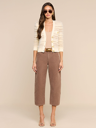 A woman wearing sunglasses poses against a plain beige background, styled in the Ariadne Cardigan—a cream, cotton-blend cardigan with mixed stitches and subtle stripes—layered over a beige top, paired with brown cropped pants and nude heels.