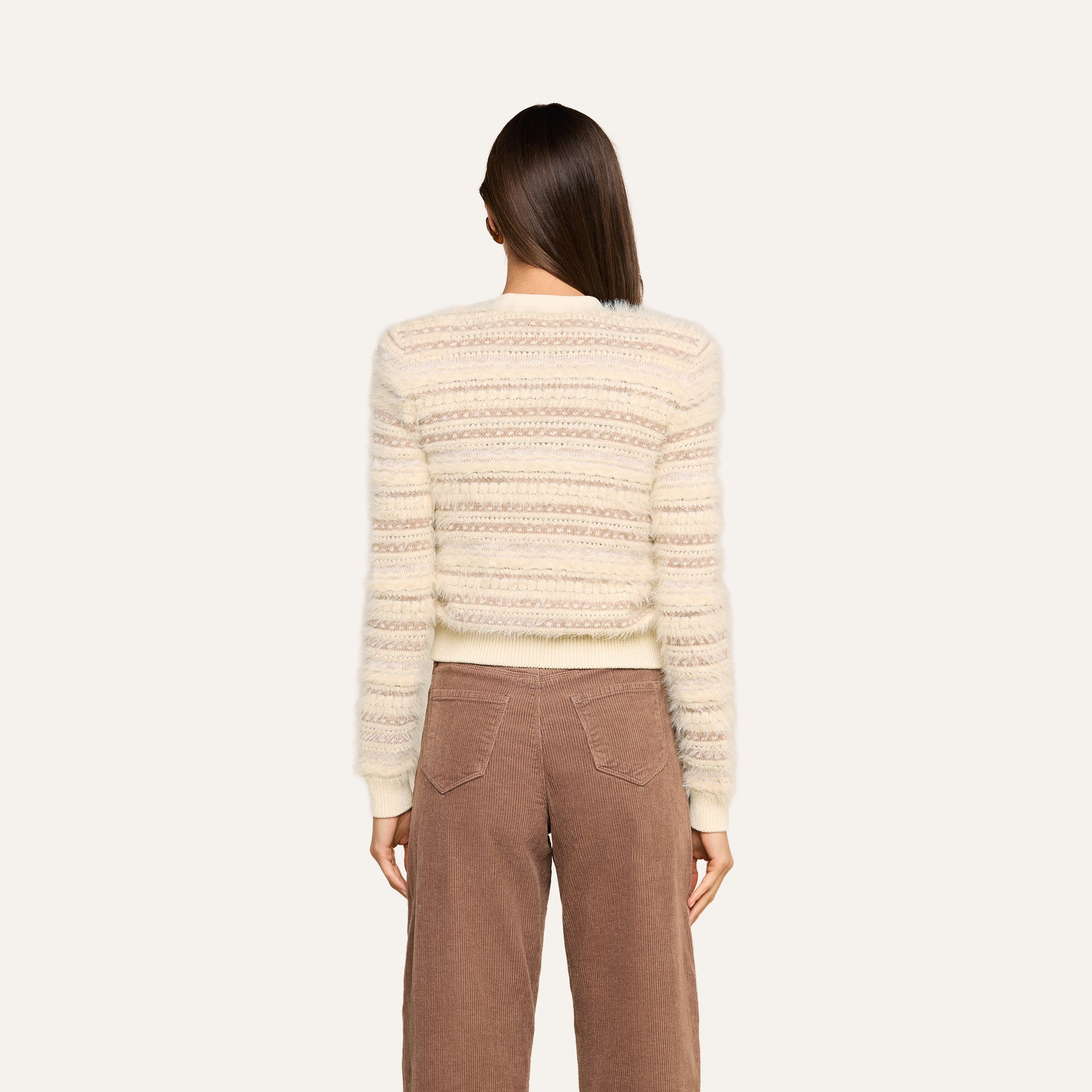 A person with straight brown hair, seen from behind, wears the Ariadne Cardigan—a cream and beige striped cotton-blend sweater—paired with high-waisted brown corduroy pants against a plain light background.