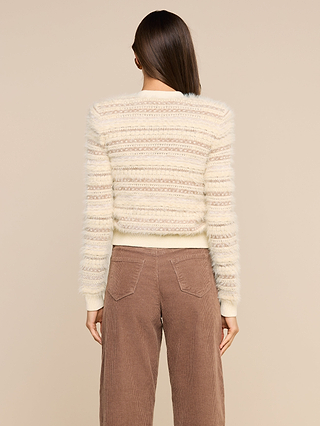 A person with straight brown hair wears the Ariadne Cardigan, a cream and beige striped fuzzy sweater crafted from cotton-blend eyelash yarns, along with brown corduroy pants, standing with their back to the camera against a beige background.