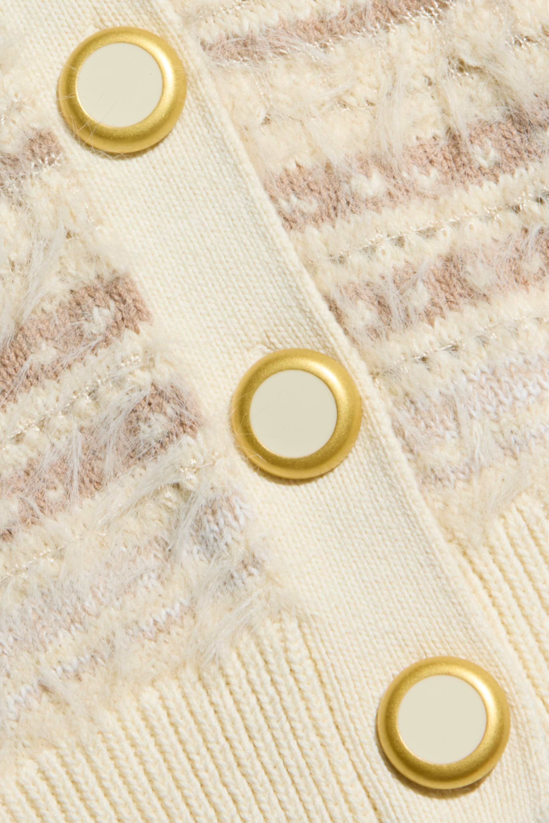 Close-up of the Ariadne Cardigan, a cream-colored knitted sweater showcasing a textured pattern created with mixed stitches and decorated with gold-rimmed round buttons down a fabric strip.