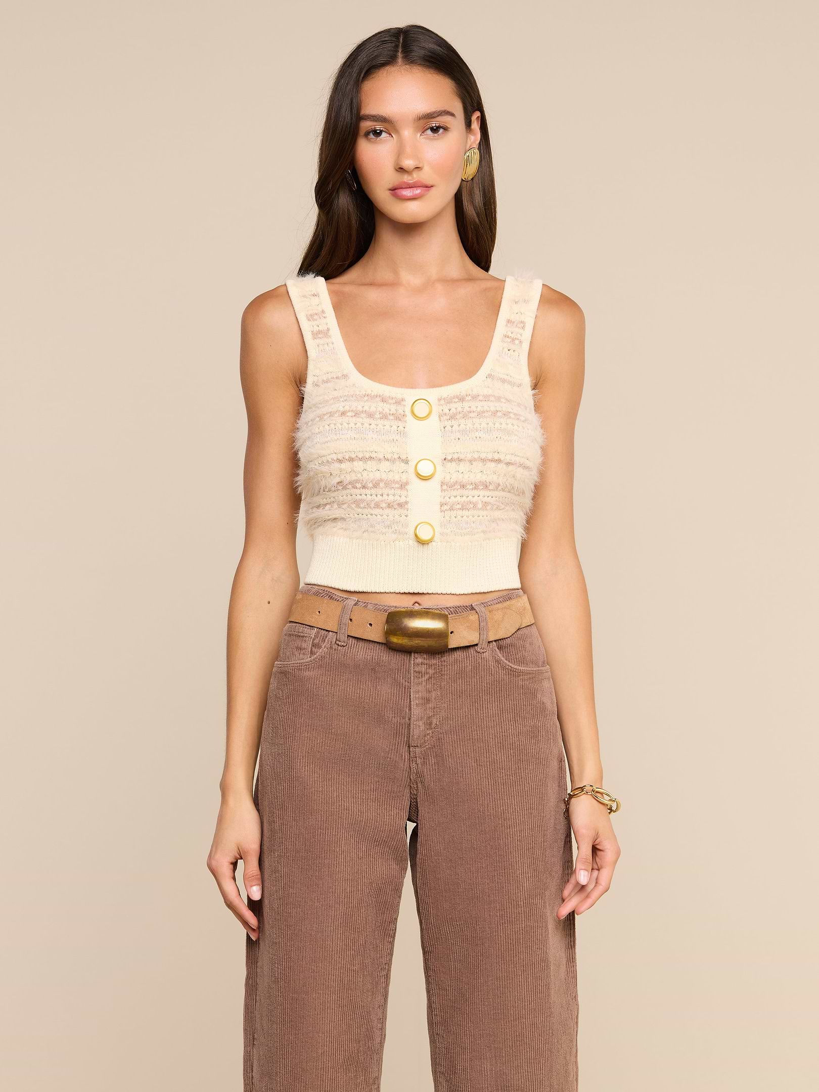A woman wears the Galene Tank, a sleeveless beige cropped knit top with three decorative buttons, paired with brown high-waisted pants, a wide belt with a brass buckle, gold earrings, and a bracelet, as she stands against an off-white background.