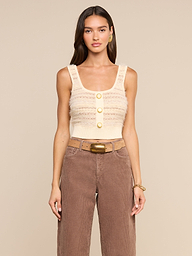 A woman wears the Galene Tank, a sleeveless beige cropped knit top with three decorative buttons, paired with brown high-waisted pants, a wide belt with a brass buckle, gold earrings, and a bracelet, as she stands against an off-white background.