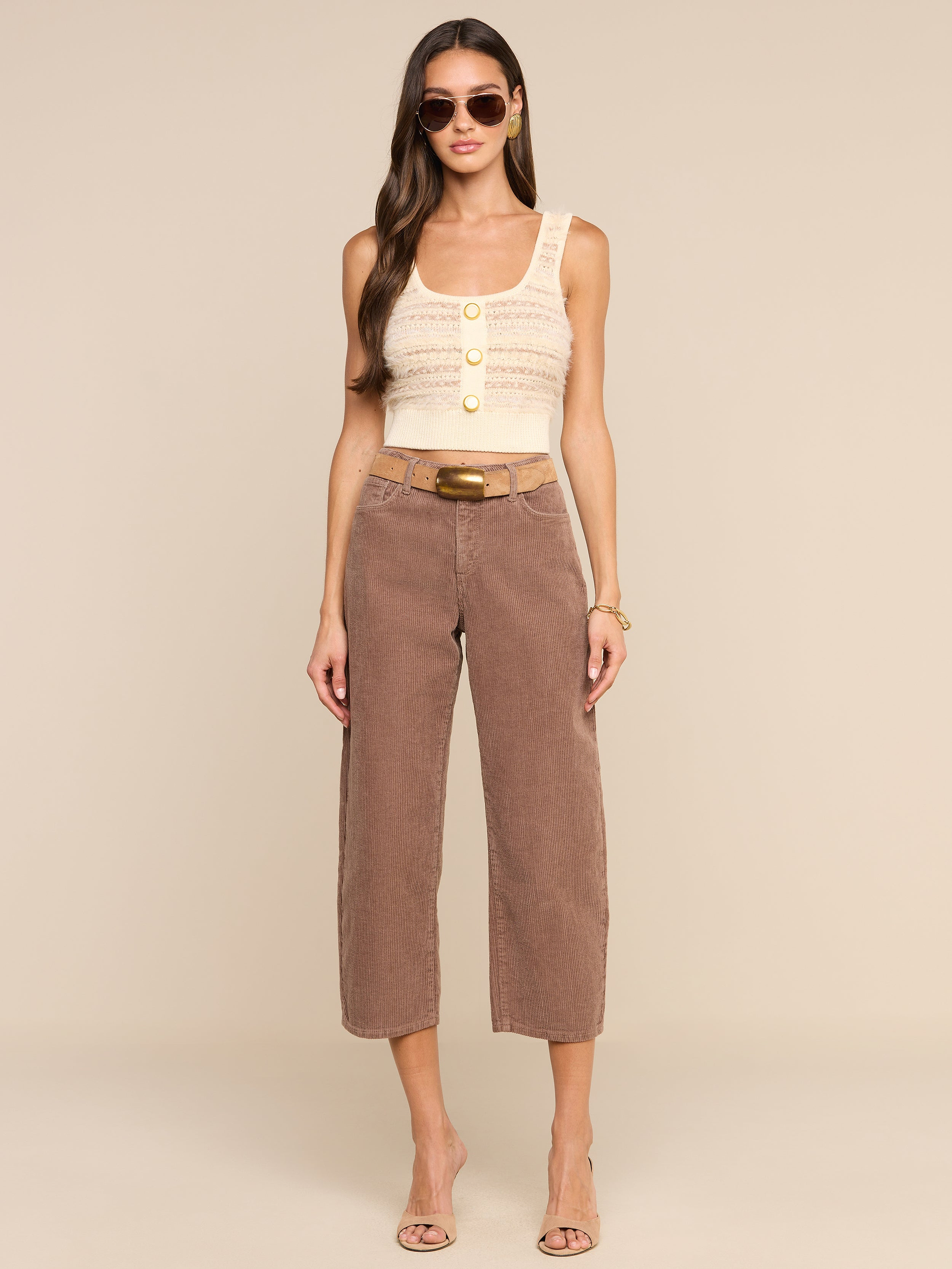 A woman in sunglasses wears the Galene Tank, a sleeveless beige top with decorative buttons, paired with brown belted cropped pants and heeled sandals, standing against a plain white background.