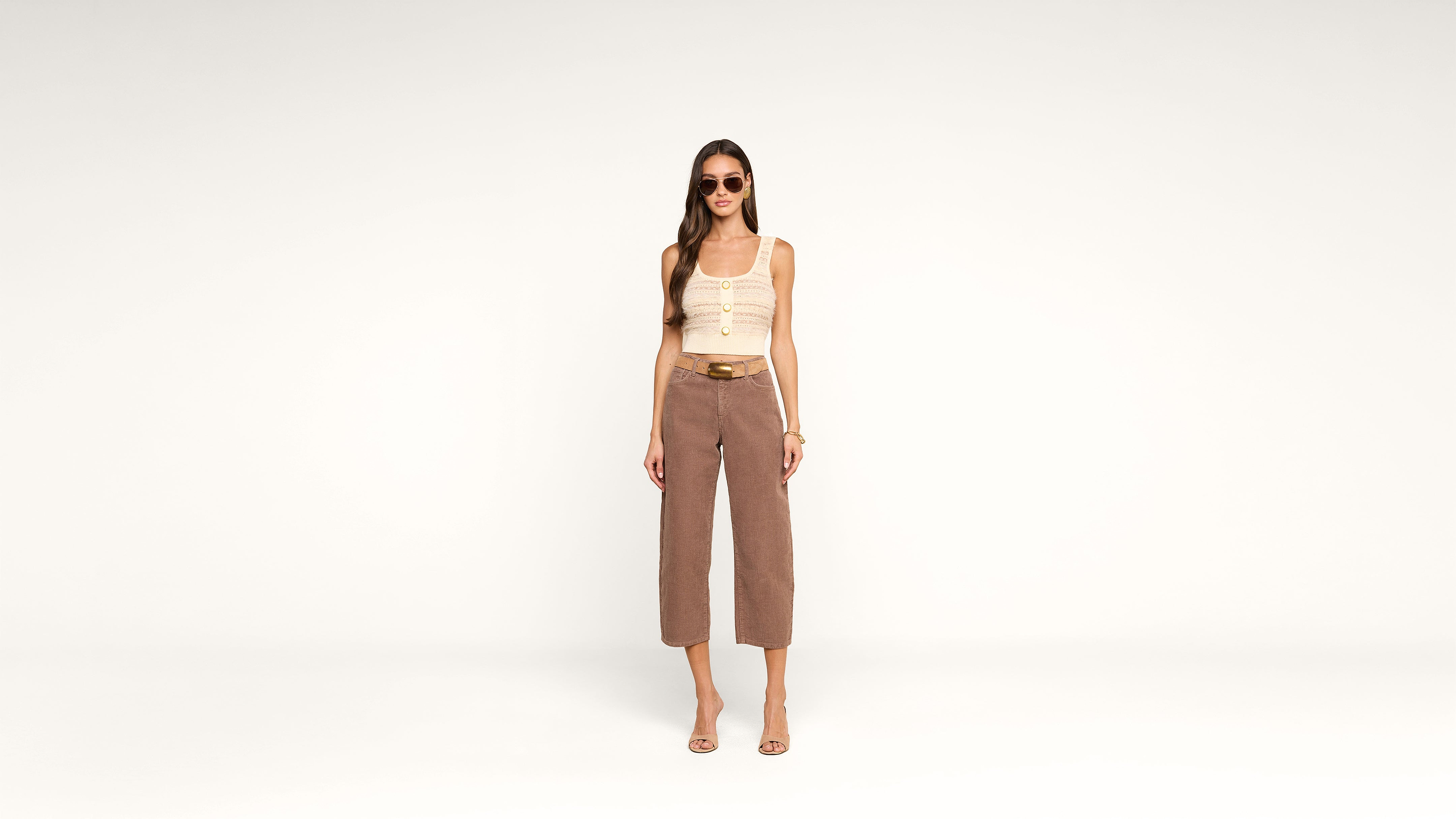 Woman wearing sunglasses, a beige sleeveless top, brown cropped pants, and heeled sandals stands confidently against a plain white background.