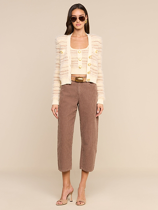 A woman stands facing forward wearing sunglasses, a cream-colored textured cardigan with vintage-style buttons, the Galene Tank—a matching cropped knit tank—taupe high-waisted pants, a gold belt, and nude heeled sandals. The background is plain beige.