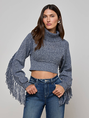 A woman wears the L'AGENCE Maddison Fringed Turtleneck, a marled blue cropped sweater with fringe on the sleeves, paired with high-waisted jeans, standing against a plain light gray background.