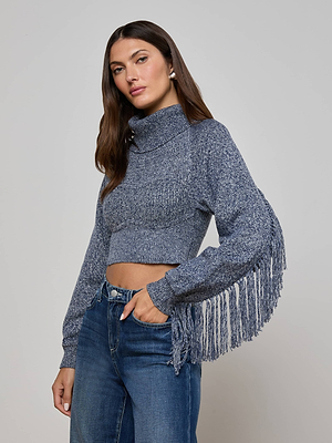 A woman in the L'AGENCE Maddison Fringed Turtleneck—a marled blue knit crop sweater with long sleeve fringe—pairs it with high-waisted blue jeans, posing with one hand in her pocket against a light gray background.
