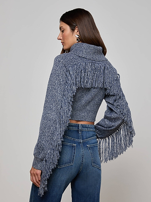 A woman with long brown hair wears the L'AGENCE Maddison Fringed Turtleneck, a marled blue statement sweater with fringe on the back and sleeves, paired with high-waisted blue jeans and large silver earrings against a plain background.