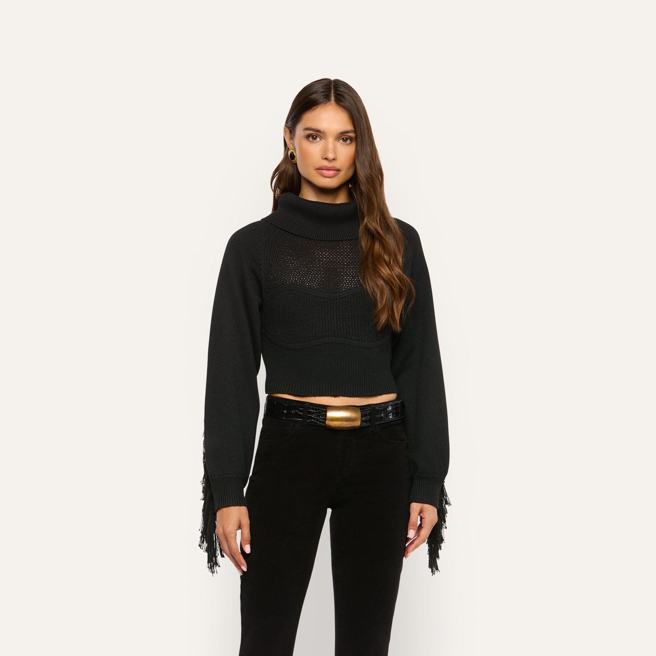 A woman wears a marled black Maddison Turtleneck featuring a cropped fit, fringe detailing on the sleeves, and a black belt with a gold buckle.